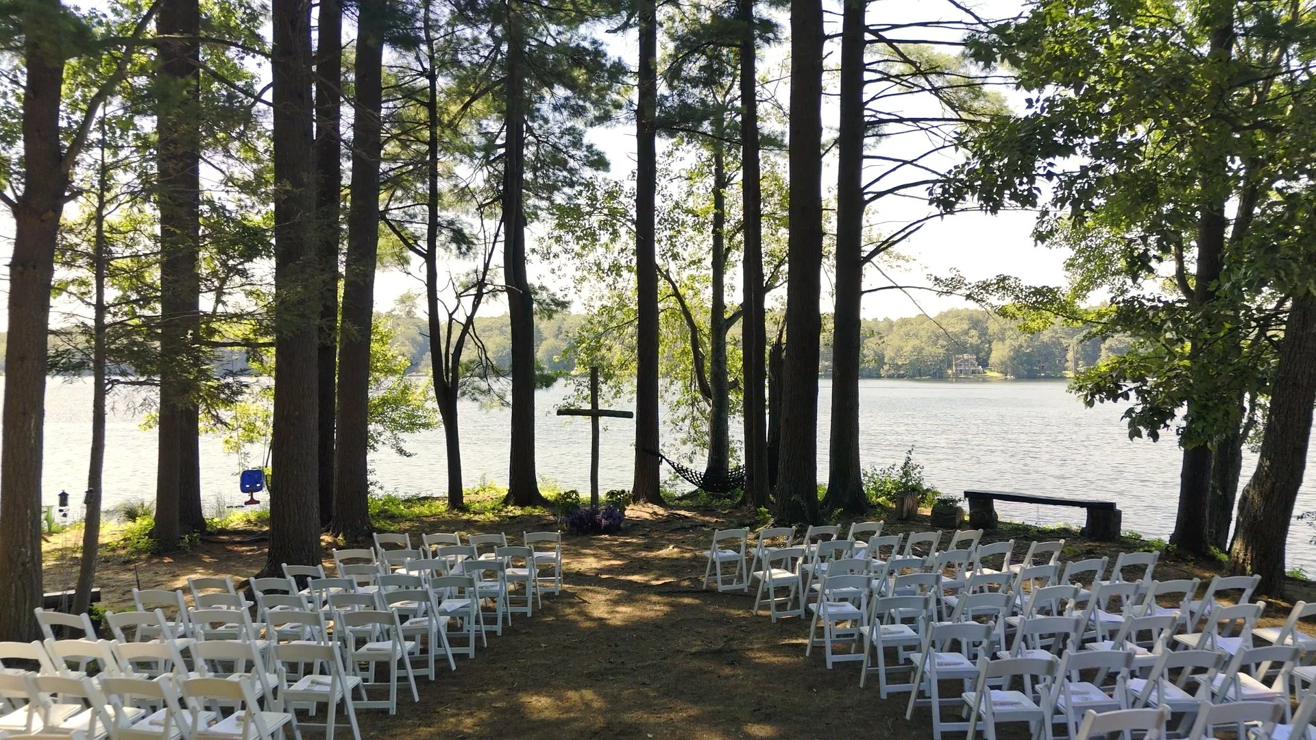Outdoor wedding setup by a lake with white chairs arranged in rows facing a wooden cross under trees, with a hammock hanging between trees, and a bench near the water.