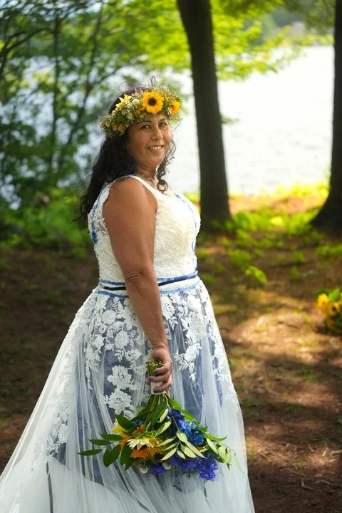 A couple in wedding attire standing close together outdoors, smiling, with the groom gently touching the bride's arm. The bride has platinum blonde hair with a floral crown, wearing a lace wedding dress, and the groom is in a black suit with a white shirt and black bow tie. The background is blurred with greenery.