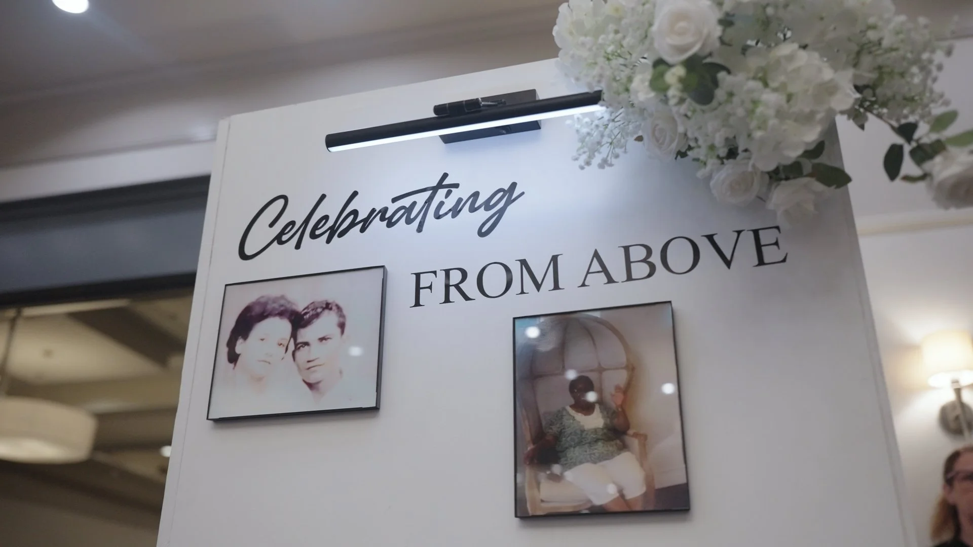 A white display board at a wedding reception with the words 'Celebrating From Above' and two photographs, one of two women and one of a woman sitting in a chair, with a bouquet of white flowers on top.