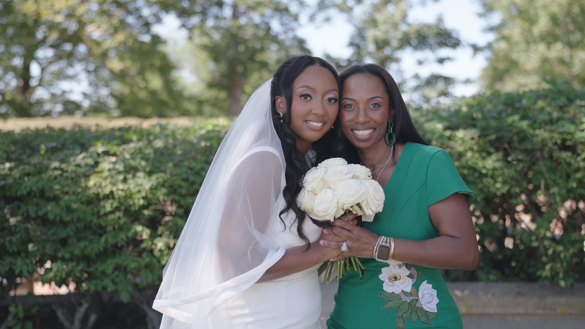 Long wooden outdoor dining table decorated with floral arrangements, candles, and wooden chairs underneath string lights, surrounded by trees.
