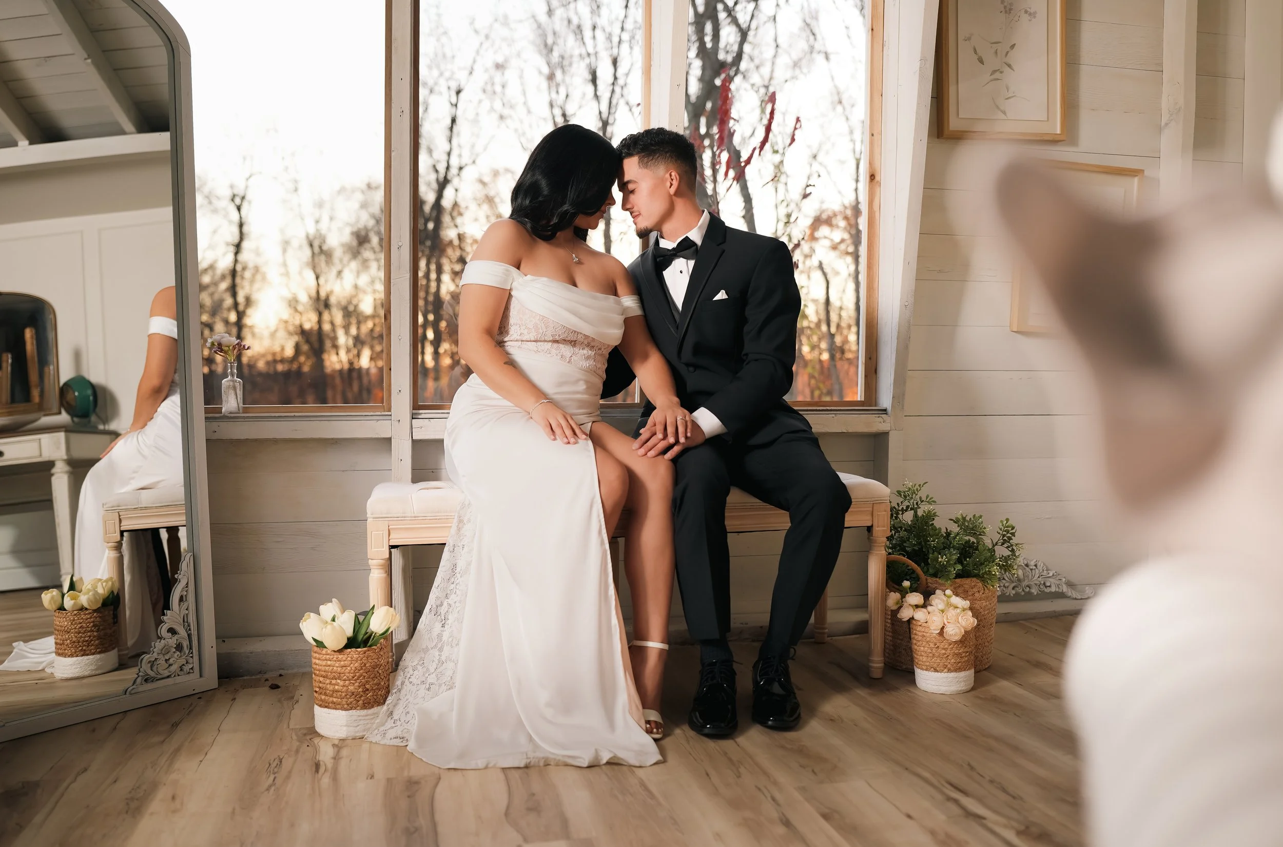 A bride in a white dress with a veil dancing joyfully while the groom, holding a bouquet of white flowers in front of his face, stands nearby against a dark wooden wall.