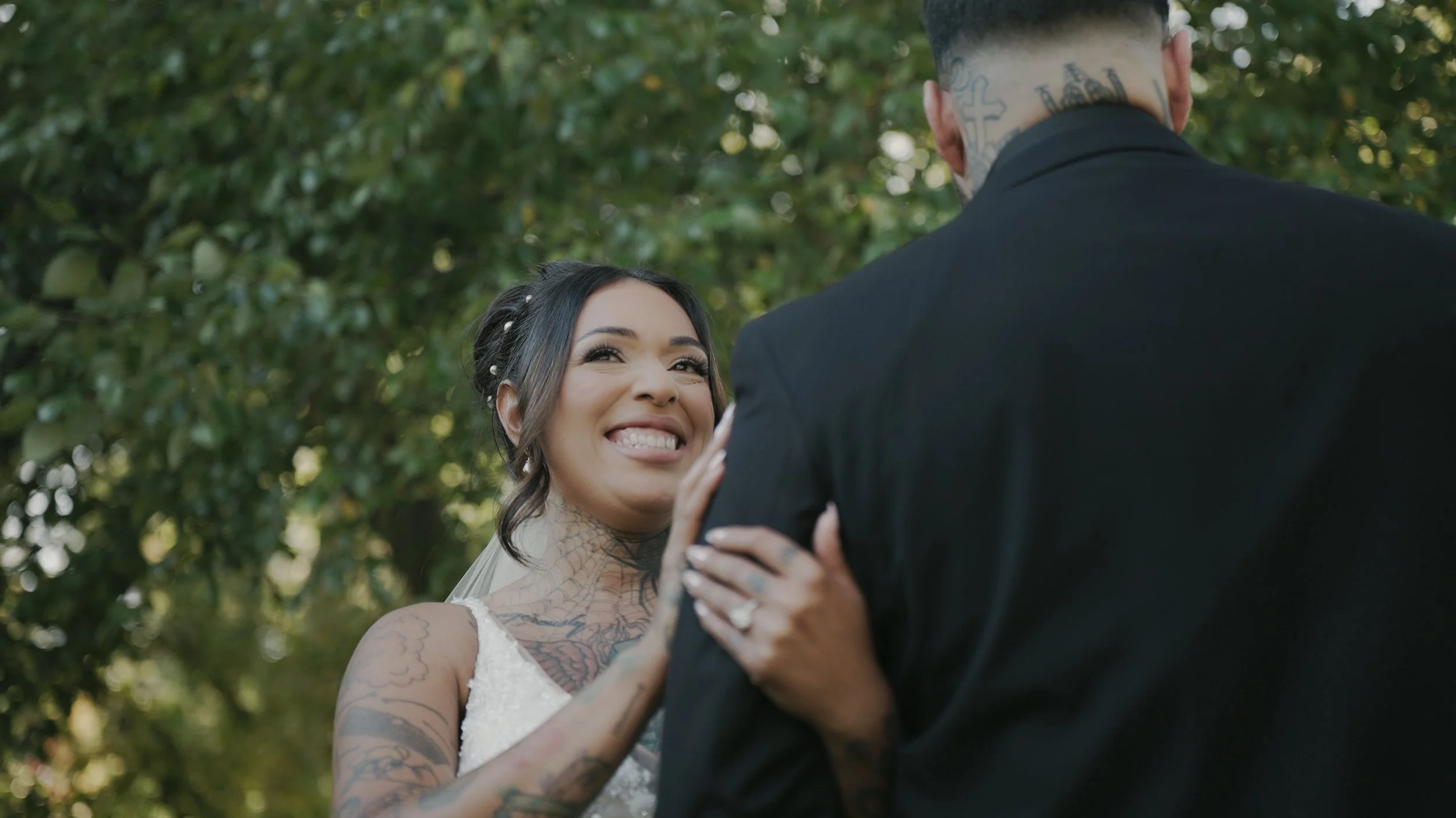 A couple in wedding attire standing close together outdoors, smiling, with the groom gently touching the bride's arm. The bride has platinum blonde hair with a floral crown, wearing a lace wedding dress, and the groom is in a black suit with a white shirt and black bow tie. The background is blurred with greenery.