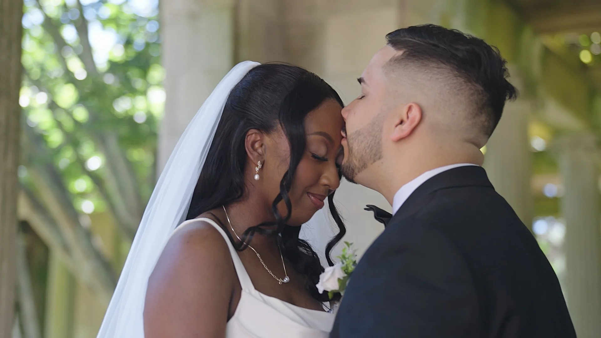 A couple in wedding attire standing close together outdoors, smiling, with the groom gently touching the bride's arm. The bride has platinum blonde hair with a floral crown, wearing a lace wedding dress, and the groom is in a black suit with a white shirt and black bow tie. The background is blurred with greenery.