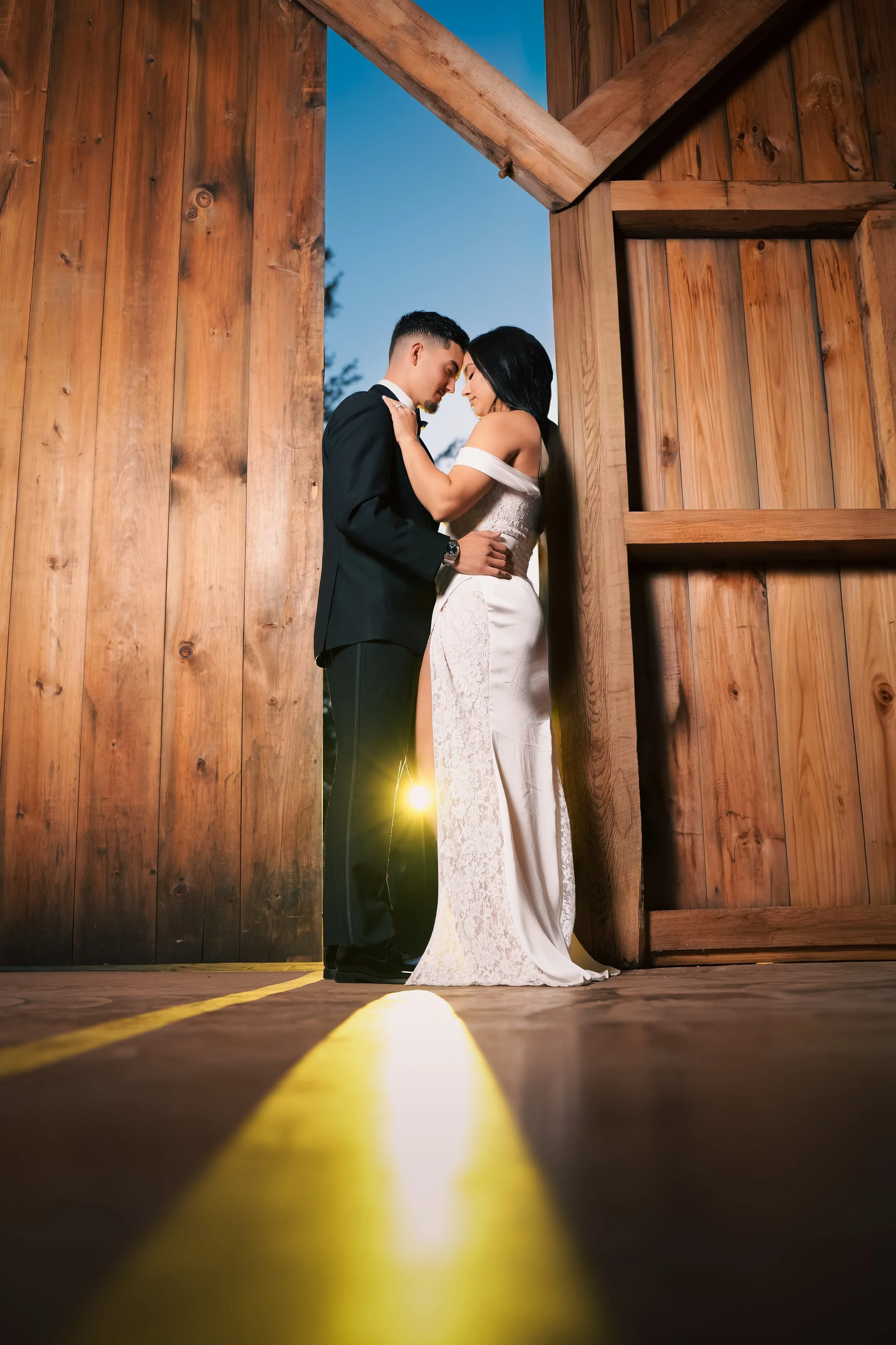 A bride in a white dress with a veil dancing joyfully while the groom, holding a bouquet of white flowers in front of his face, stands nearby against a dark wooden wall.