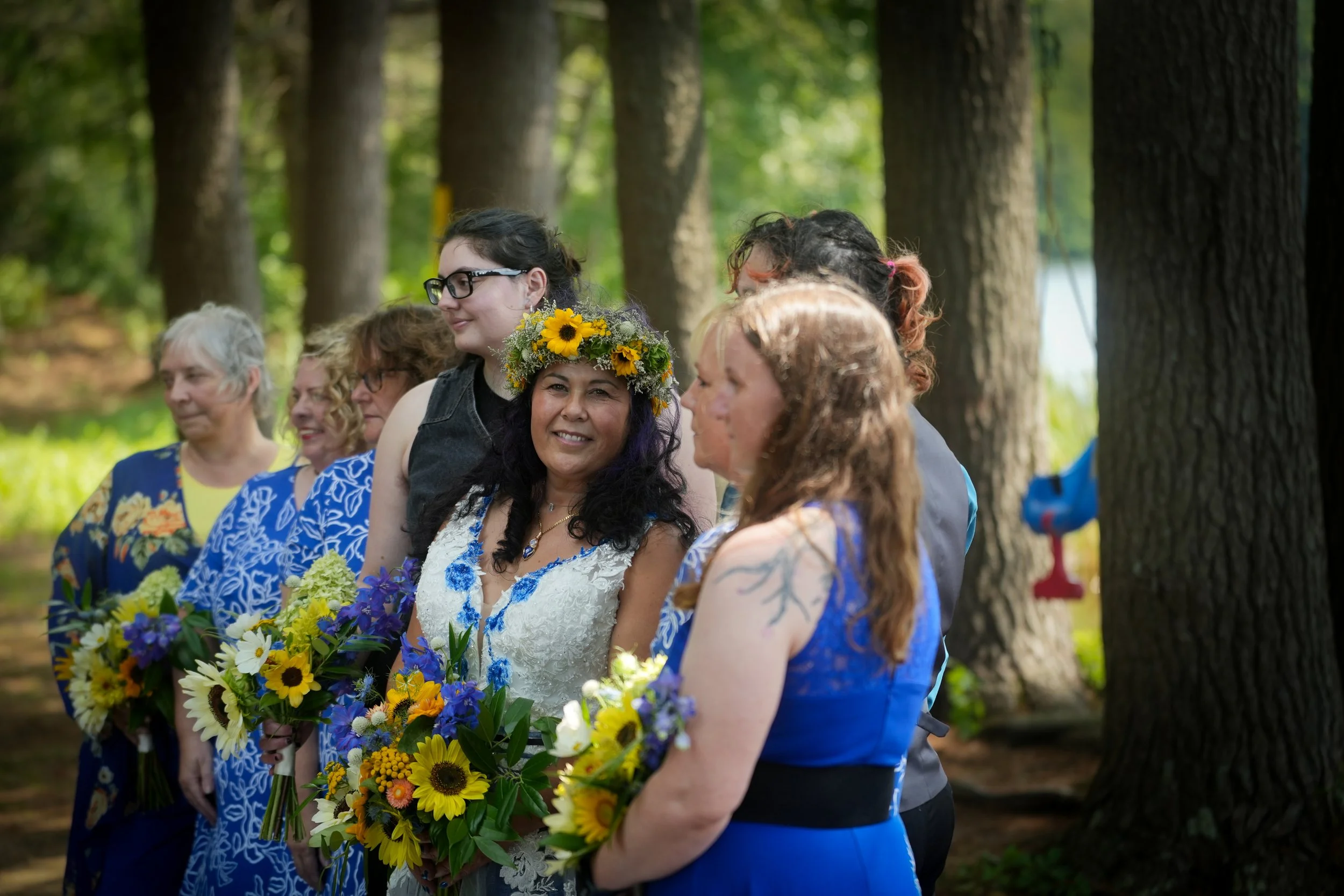 A group of women dressed in blue and one in a wedding dress with a sunflower crown, holding bouquets, standing outdoors in a wooded area during a celebration or ceremony.