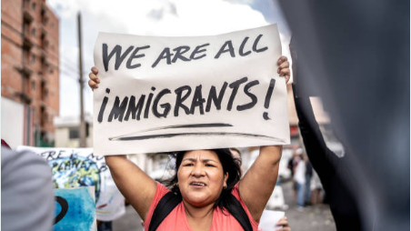 A woman holding a sign that reads 'WE ARE ALL IMMIGRANTS!' at a protest or rally.