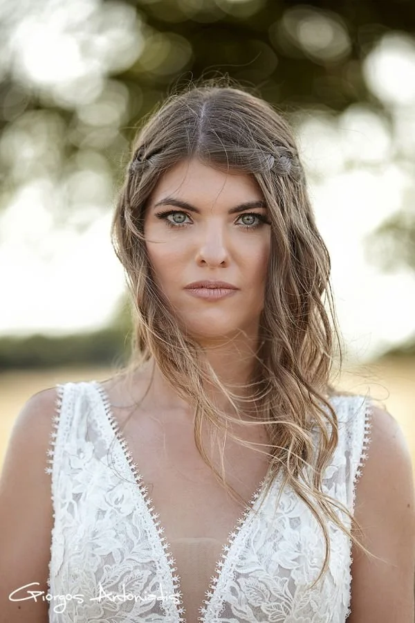 A young woman with long wavy brown hair and blue eyes, wearing a white lace dress, standing outdoors with blurred trees in the background.