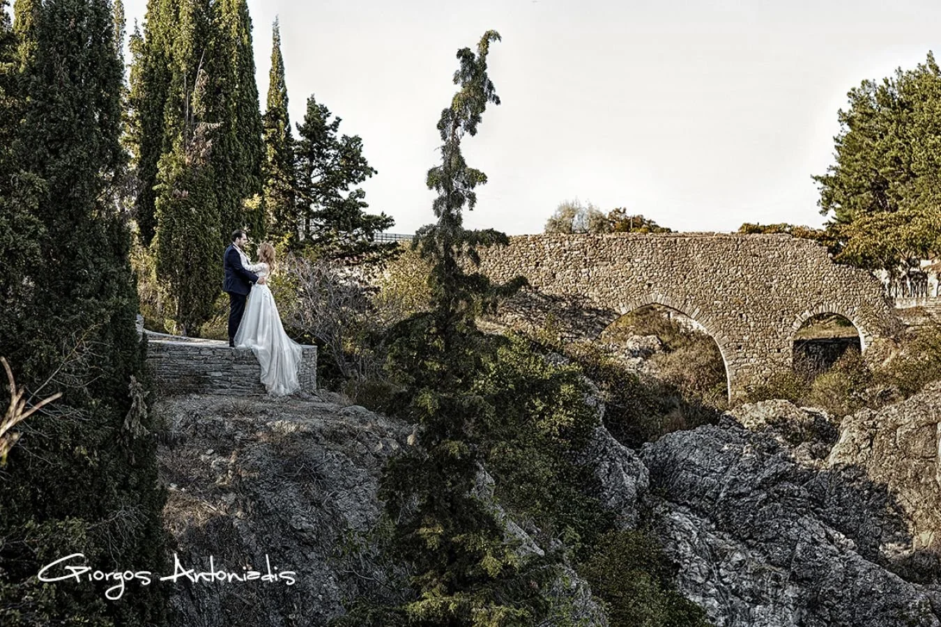 A bride and groom holding each other on a rocky ledge amidst tall trees, with an ancient stone bridge in the background.