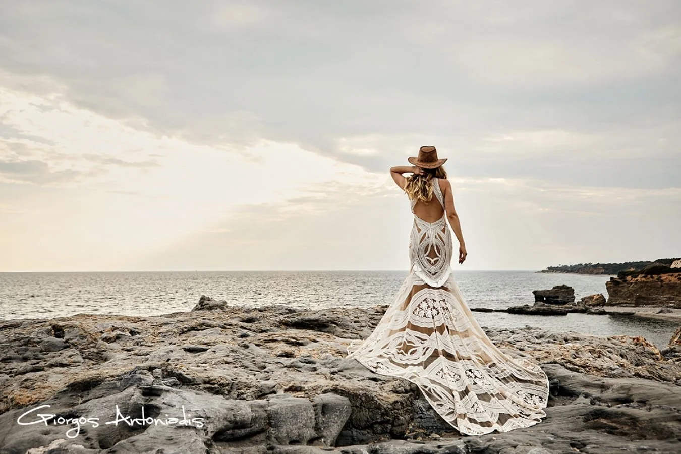A woman in a white lace gown and wide-brimmed hat standing on rocky shoreline facing the ocean during sunset.