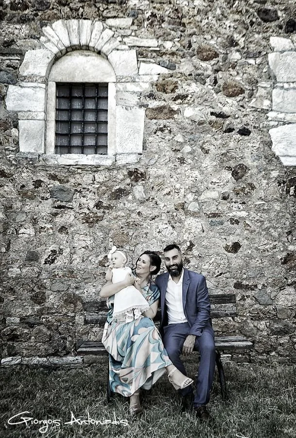 A woman, a little girl, and a man sitting on a bench against a rustic stone wall with a small barred window. The woman is holding the girl, and the man is smiling. The woman is in a colorful dress, and the man is in a suit.