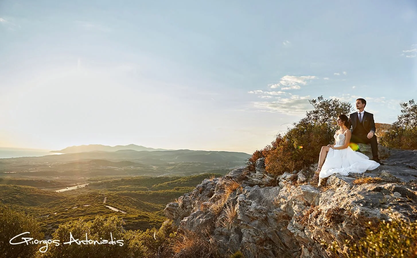 A bride in a white wedding dress sitting on rocky terrain with a groom in a black suit standing behind her, overlooking a landscape of hills and valleys during sunset.