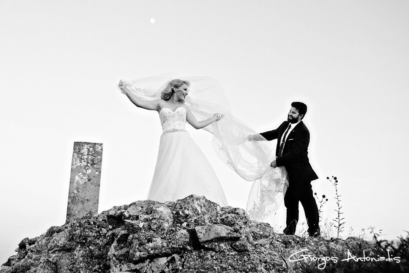 A black and white photo of a bride and groom standing on rocks outdoors. The bride is lifting her veil, smiling, while the groom holds her veil and smiles back.