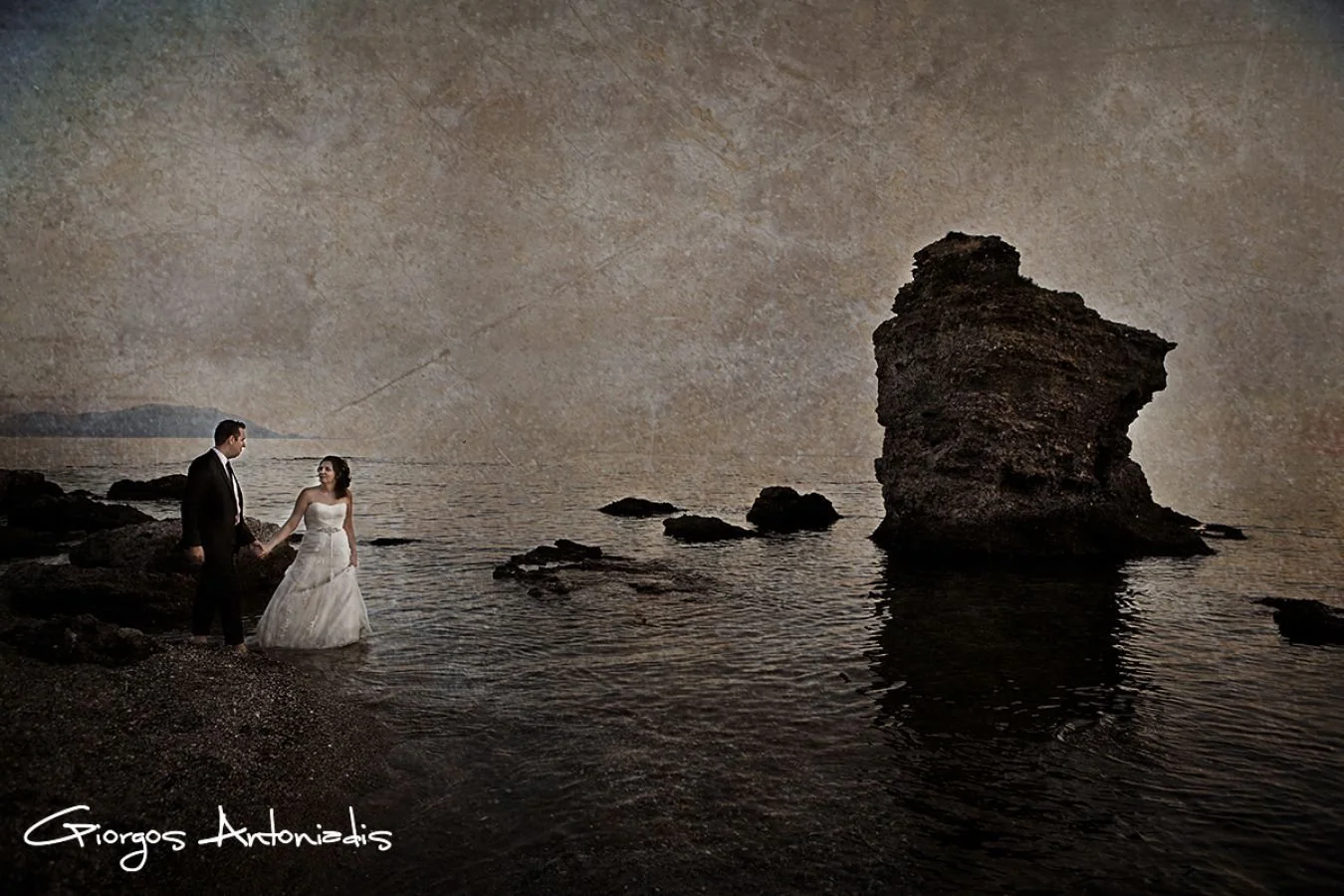A bride and groom holding hands by the water's edge on a rocky beach at sunset, with a large rock formation in the background.