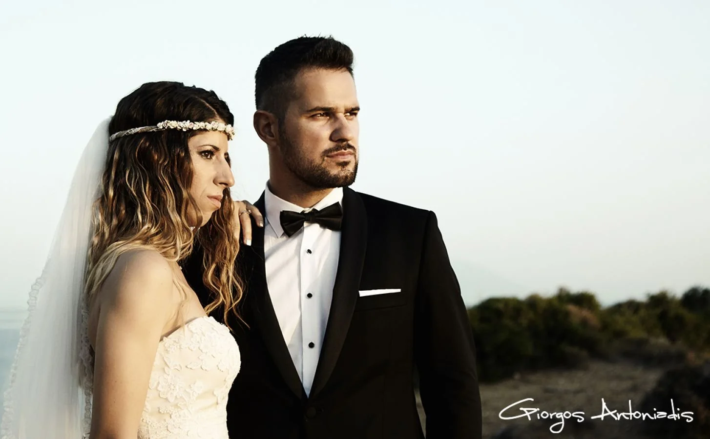 A bride and groom in wedding attire standing outdoors, gazing into the distance with a clear sky and some greenery in the background.