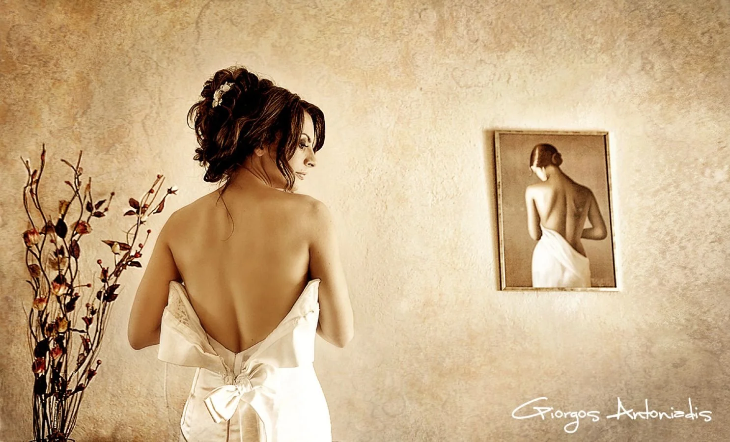 A woman with dark hair styled with a floral hair accessory is sitting on a bed, facing a mirror on the wall, wrapped in a white satin robe with a large bow at the back, in a warmly lit room with textured walls and a decorative vase with branches near