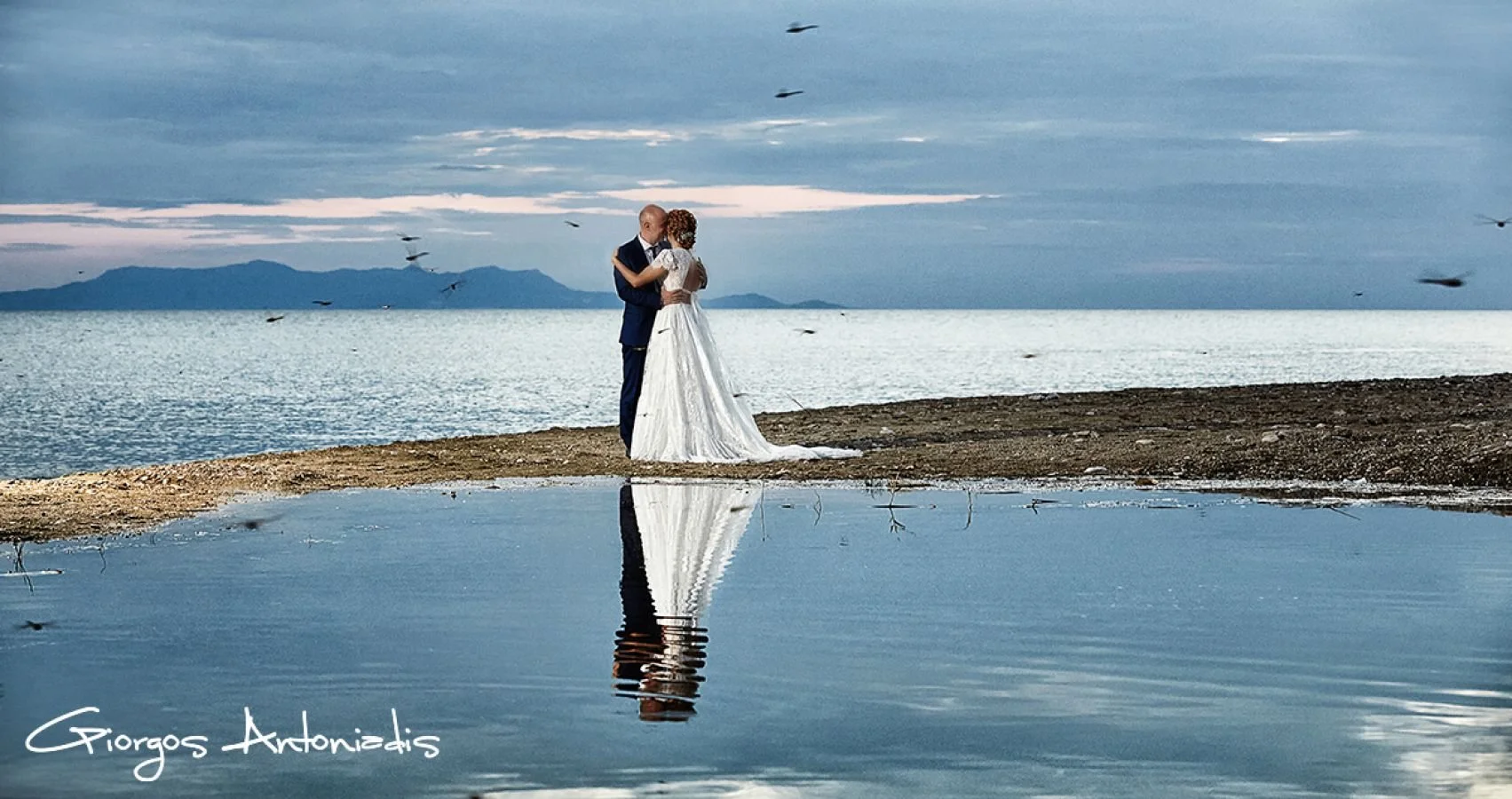 A wedding couple in formal attire embracing on a beach with a calm sea, distant mountains, and a cloudy sky in the background. Their reflection is visible in a still pool of water at their feet.