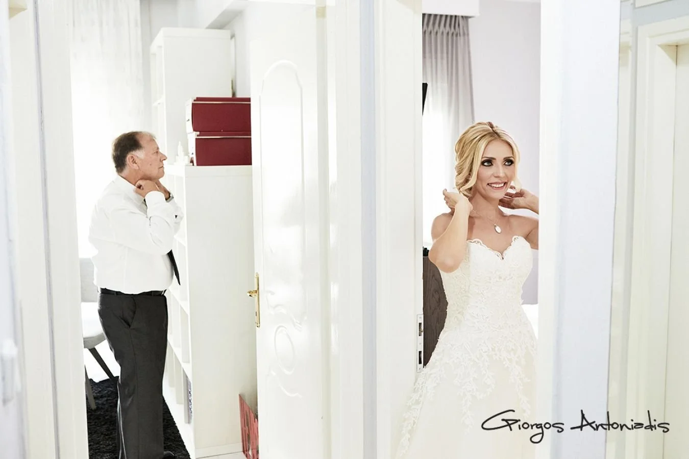 A bride in a white wedding dress adjusting her hair as an older man watches from a doorway in a bright room.
