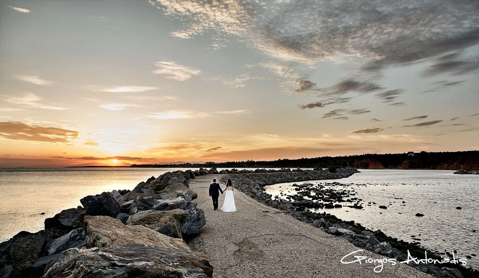 A couple dressed in formal attire, holding hands, walking along a rocky pathway by the water during sunset.