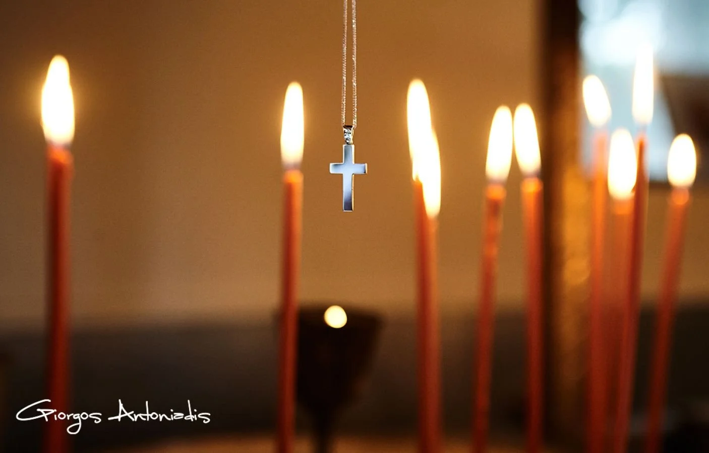 A Christian cross necklace hanging in front of lit candles on a candlestick holder.