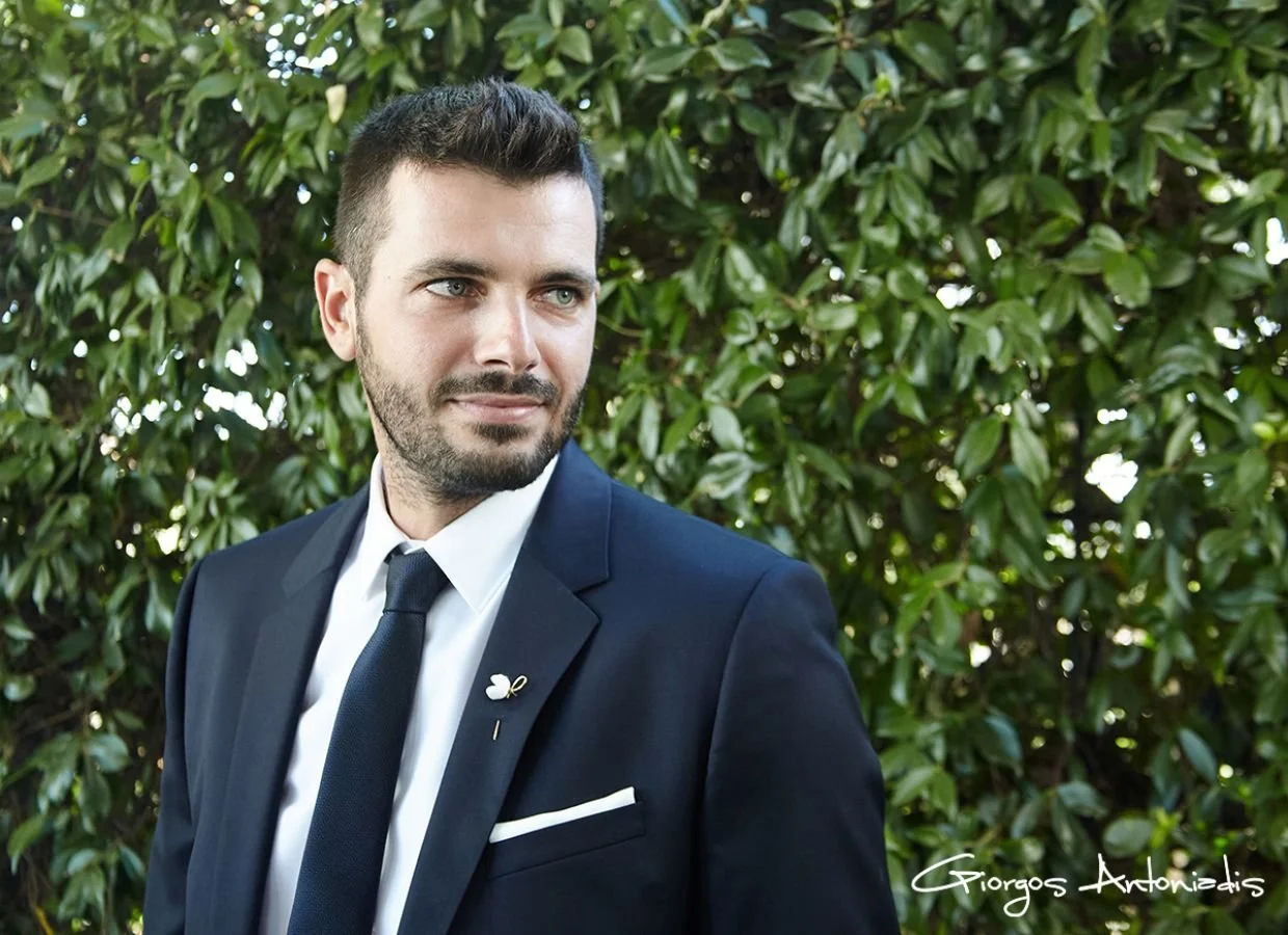 A man in a navy blue suit, white shirt, and navy tie standing outdoors in front of green foliage, looking to his right.