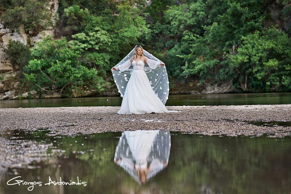 Bride in a white wedding gown stands on a pebble beach near water with greenery and trees in the background, holding her veil out.