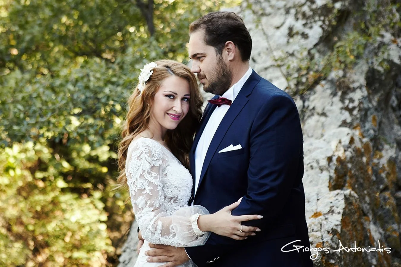 A bride and groom embracing outdoors, with the bride smiling and wearing a white lace wedding dress and the groom in a navy suit with a red bow tie, in front of a natural rocky background.