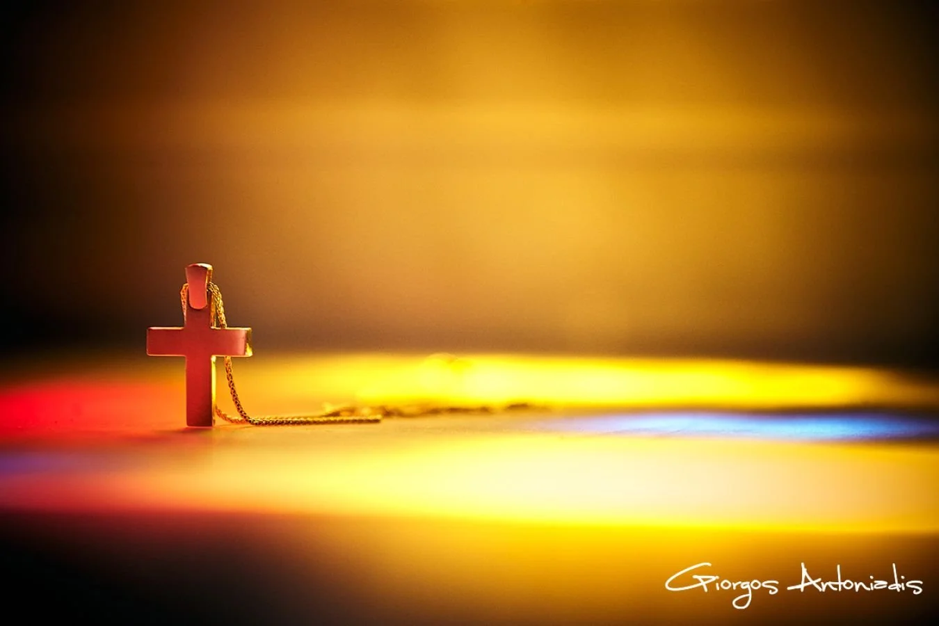 A red cross pendant with a gold chain lying on a surface illuminated with multicolored lights, creating a vibrant background.