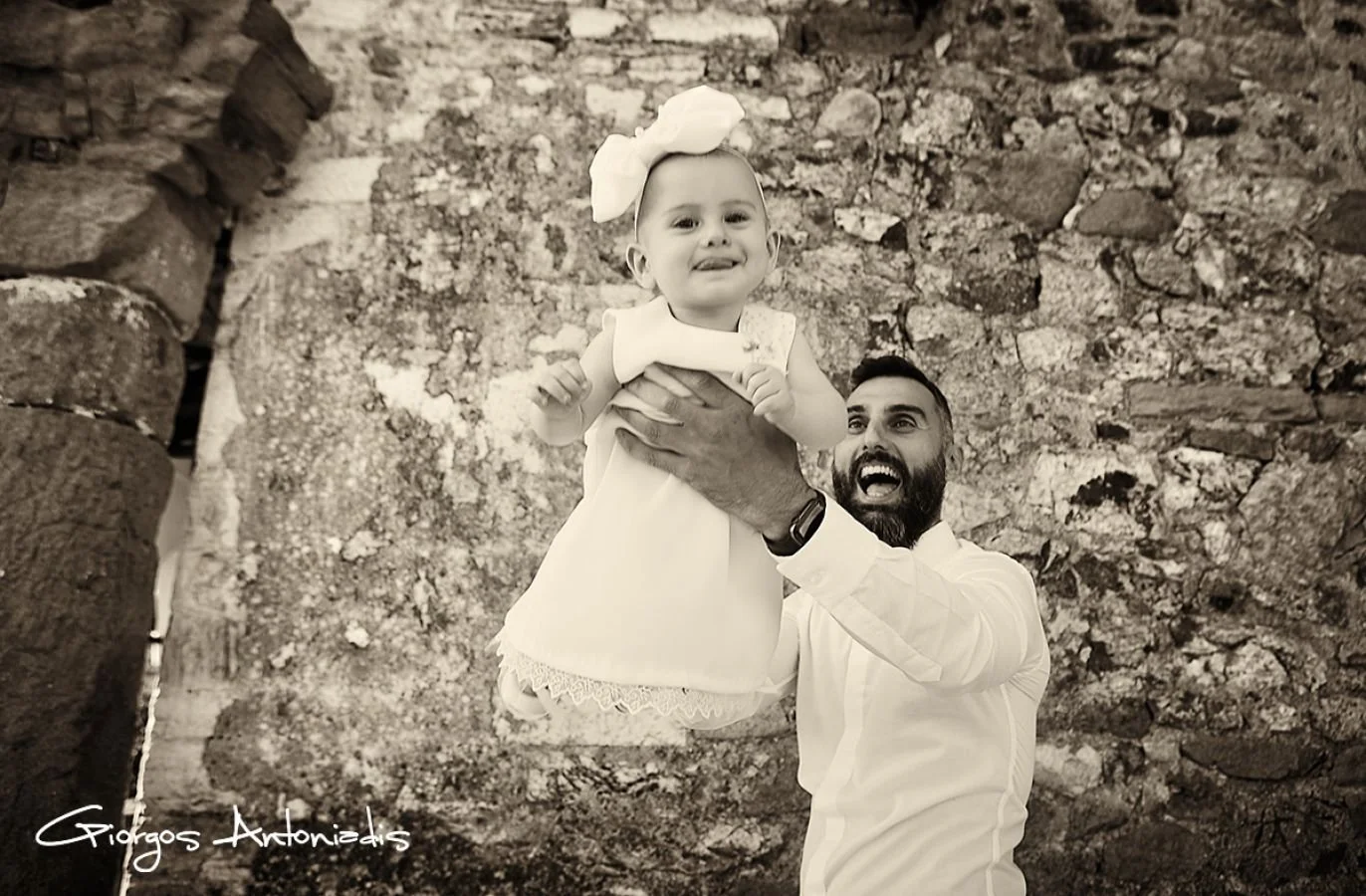 A smiling man holding a young girl in the air against a stone wall background.