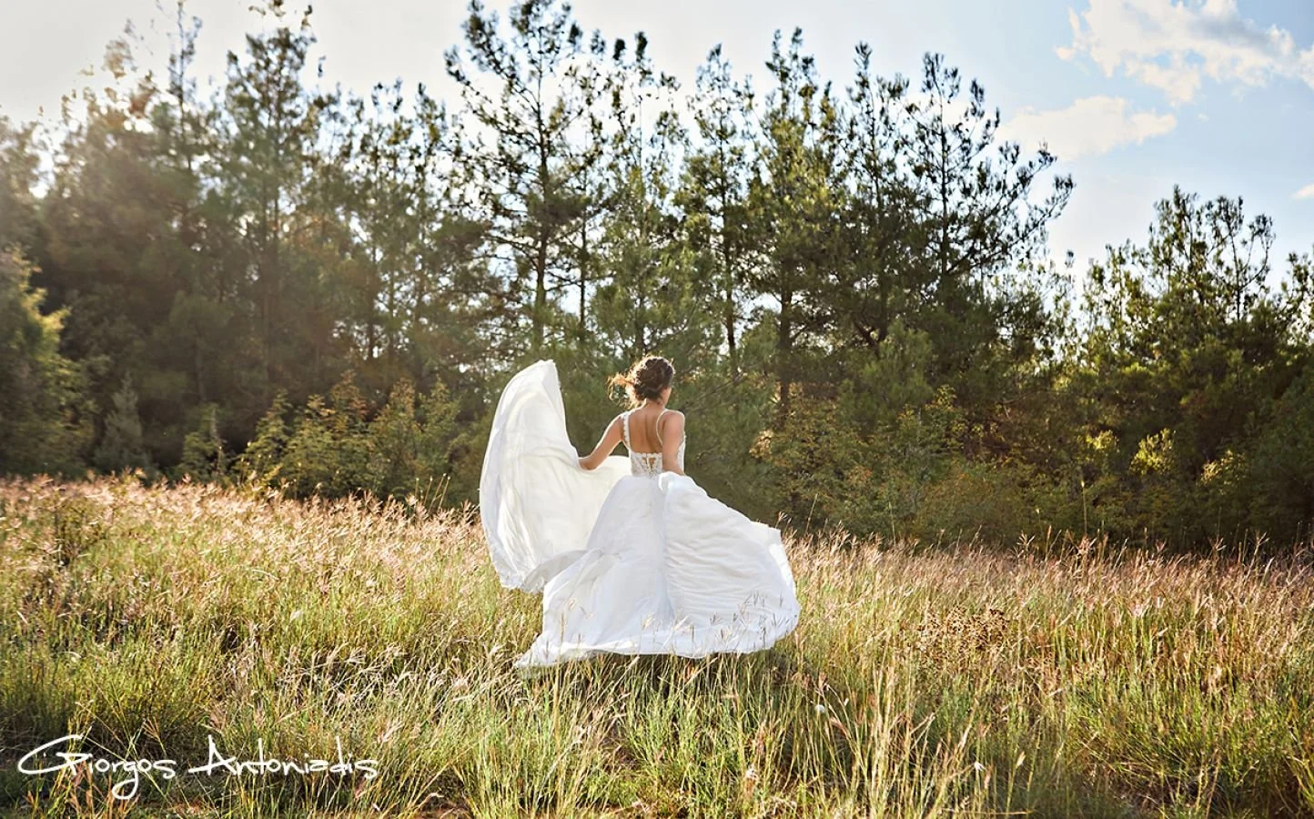 A woman in a white wedding dress walking through a grassy field with trees in the background, illuminated by sunlight.