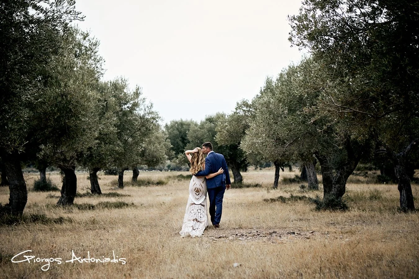 A bride and groom walk hand-in-hand through a field flanked by trees, with the bride in a white lace dress and the groom in a blue suit, embracing in a romantic moment.