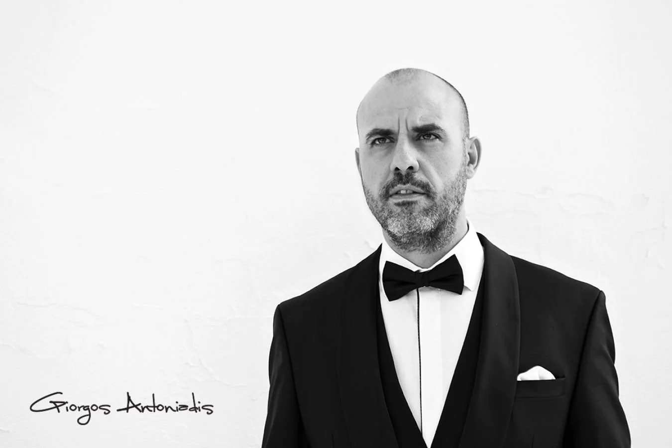 Black and white portrait of a man in a tuxedo with a bow tie, date with a serious expression, signature 'Giorgos Antonidis' at the bottom left.