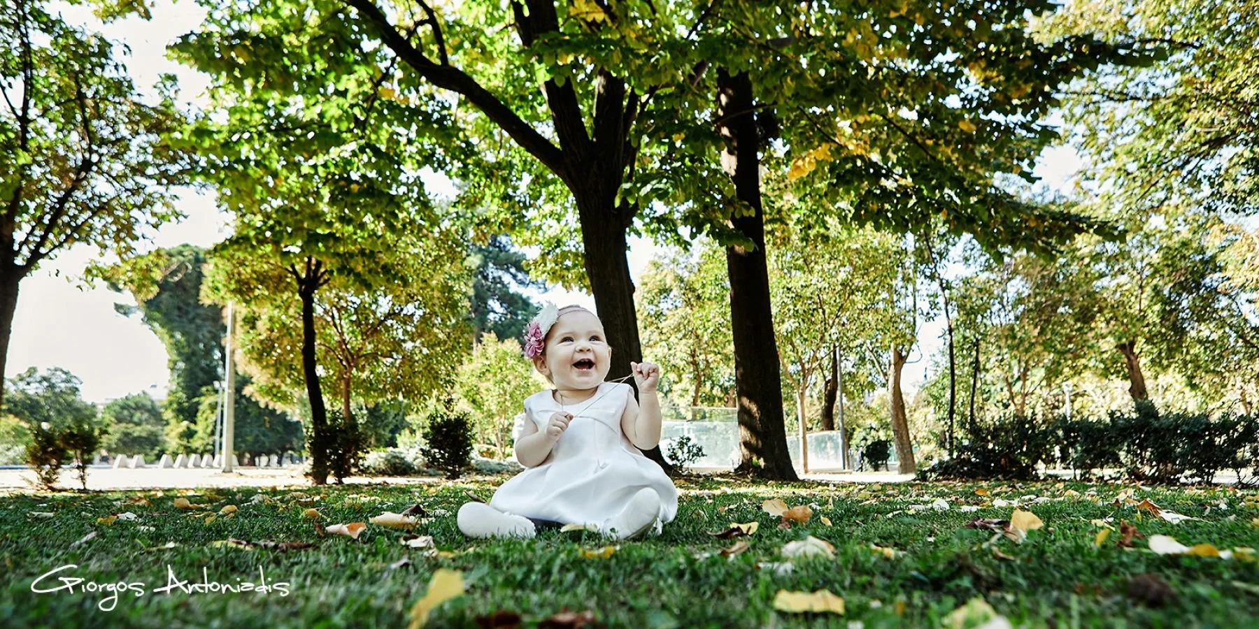 A young girl in a white dress and headband with flowers, sitting on grass in a park, smiling and holding a string, with tall trees and green foliage in the background.