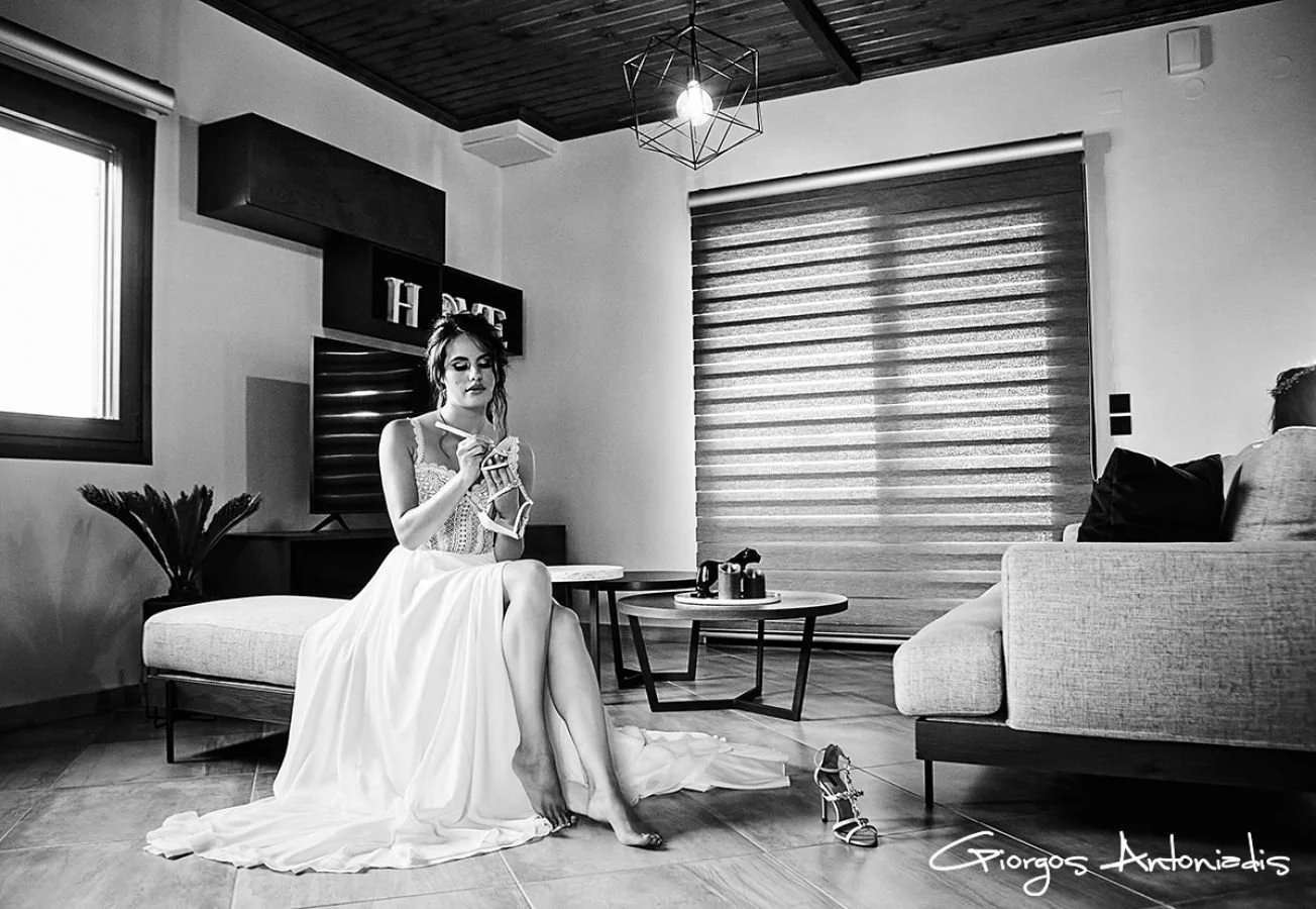 A woman in a wedding dress sitting on a couch in a modern living room, holding a phone, with a high heel shoe on the floor nearby. The room has a window, coffee tables, a sofa, and decorative items, all in black and white.