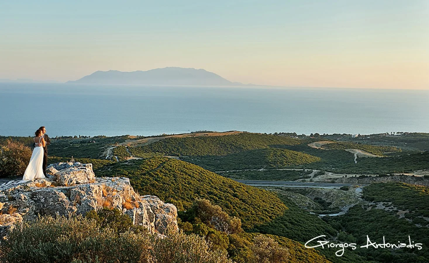 A couple dressed in wedding attire standing on a rocky hilltop overlooking lush green hills, the ocean, and a distant island during sunset.