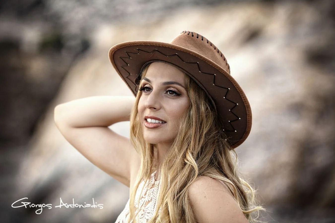 A woman with long wavy hair wearing a brown wide-brimmed hat and a sleeveless top, standing outdoors with a rocky background.