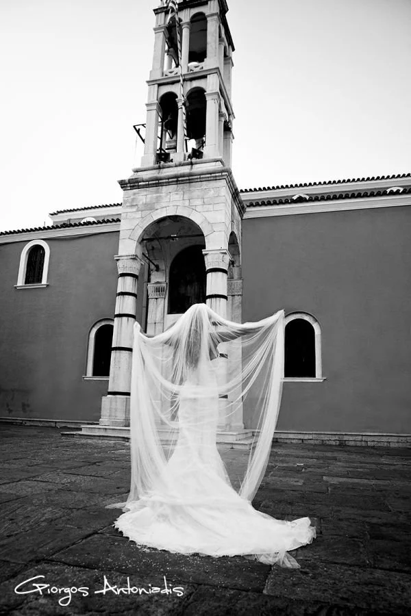 A bride in a wedding dress with a long train and veil stands with her back to the camera, holding her veil outstretched in front of a church with a bell tower in black and white.