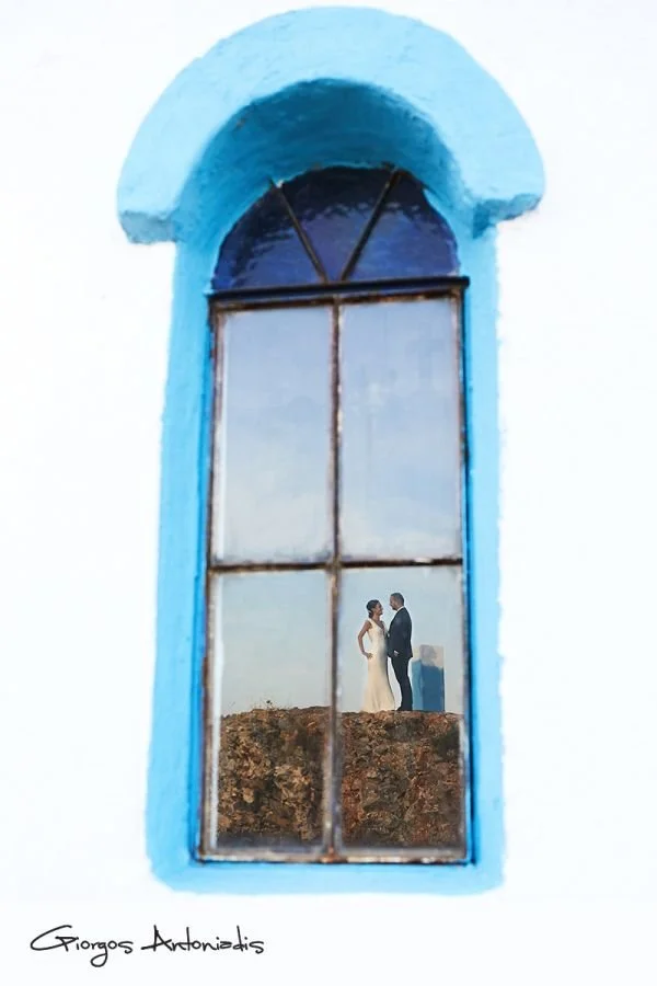 A wedding couple standing on a rocky surface seen through an arched, weathered window with blue trim.
