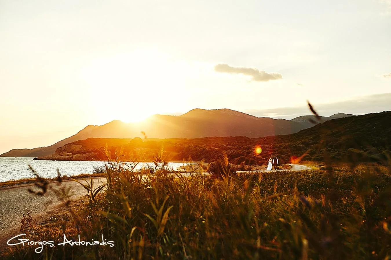 A couple dressed in wedding attire walking along a road beside a body of water during sunset, with a mountainous landscape in the background.