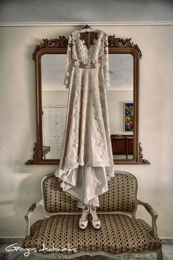 A wedding dress hangs on a hanger in front of a large ornate mirror, placed above a vintage upholstered bench. The dress features lace details, long sleeves, and a train that drapes onto the bench. A pair of white high-heeled shoes is positioned bene