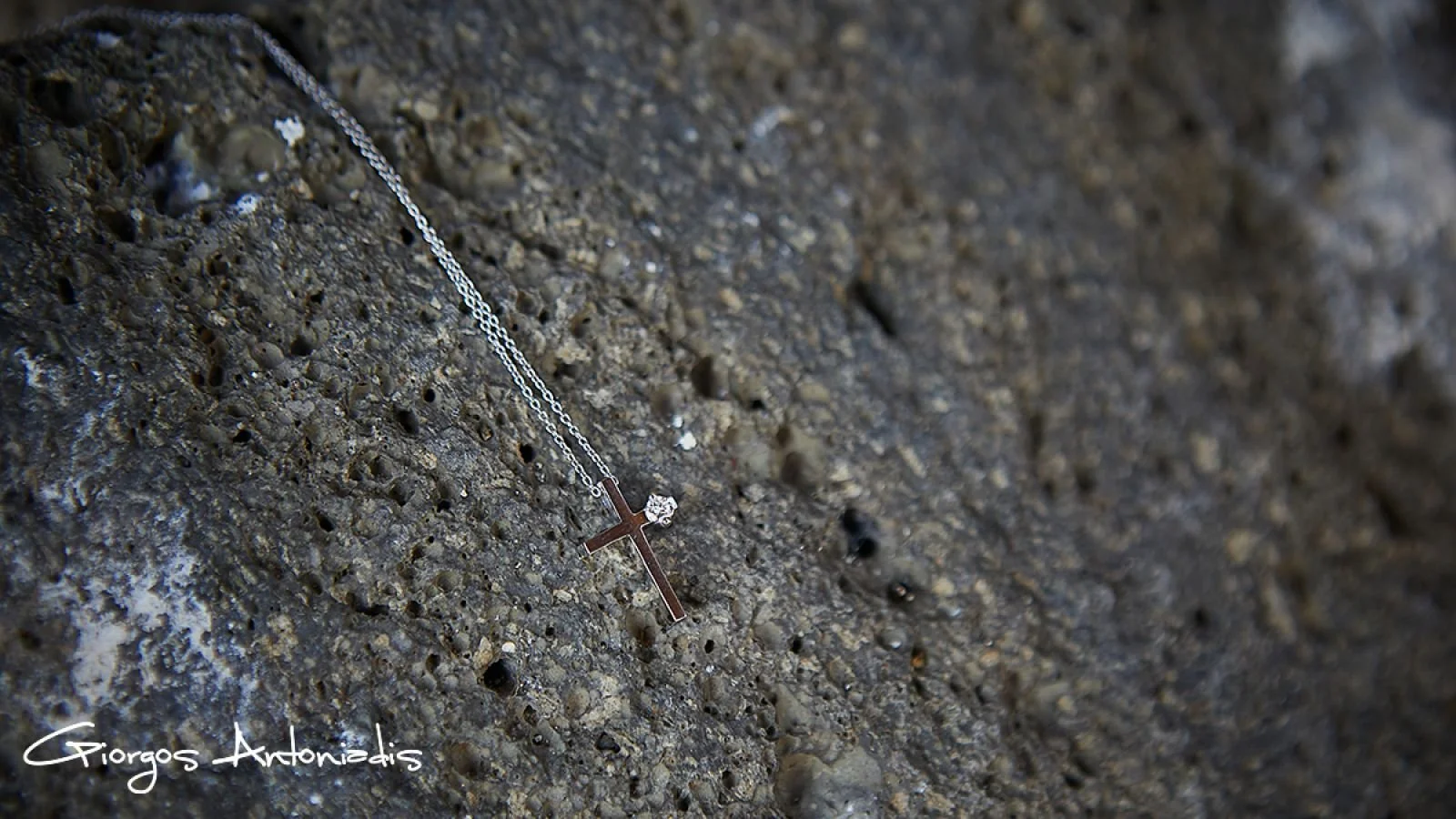 Silver necklace with a cross pendant and a small gemstone, resting on a rough, textured stone surface.