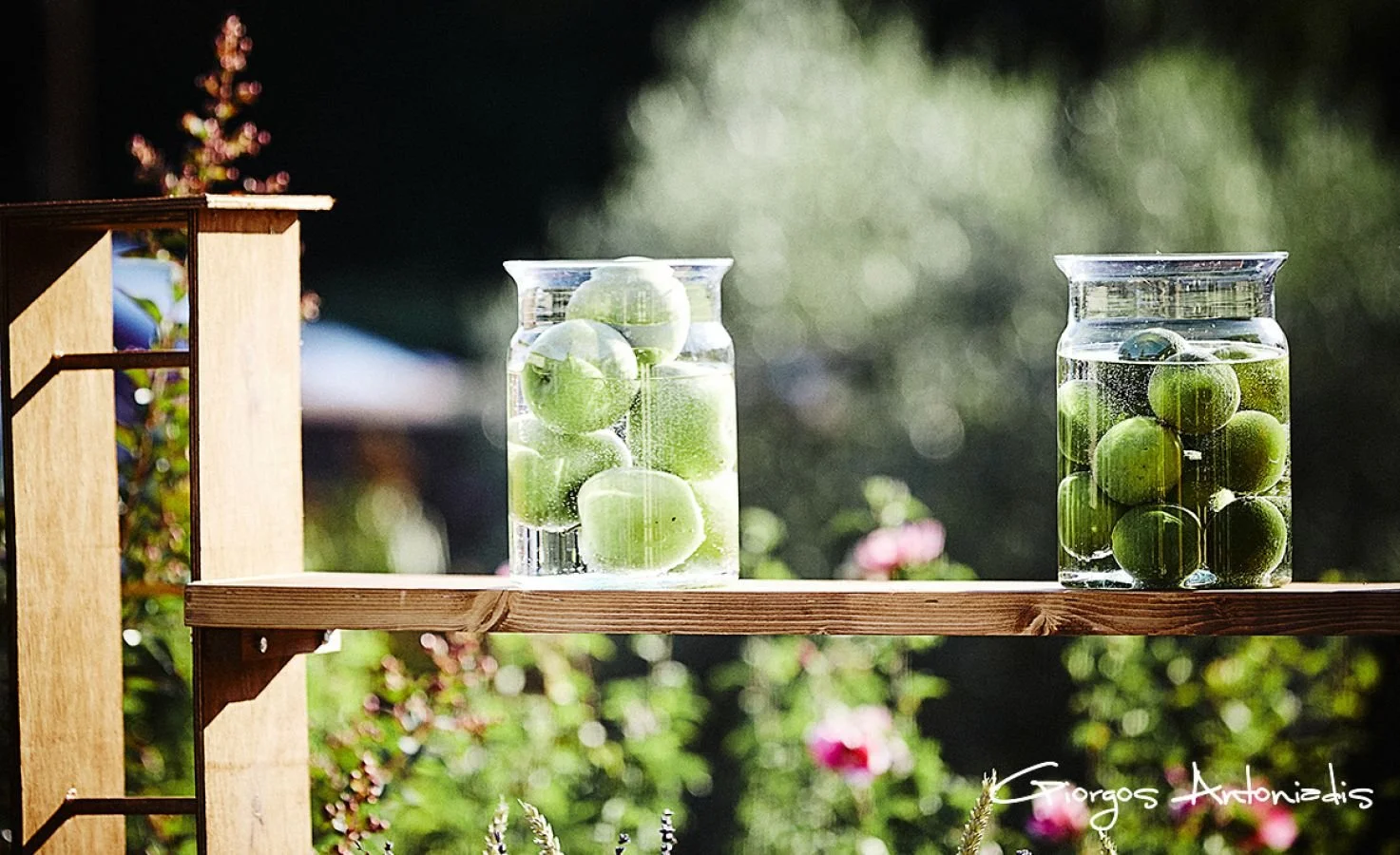 Two glass jars filled with green apples submerged in water, placed on a wooden plank outdoors during daytime.