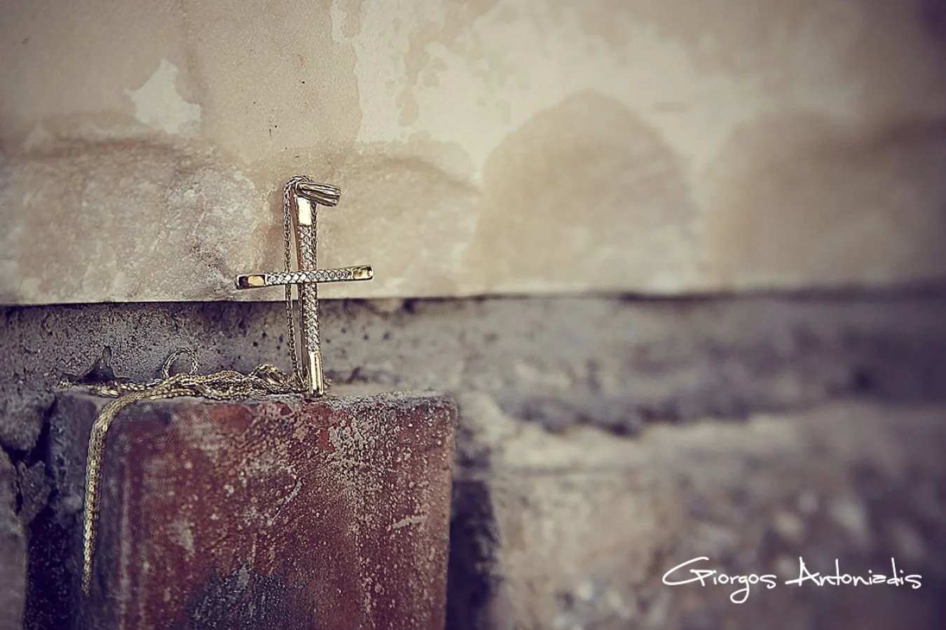 A gold necklace with a cross pendant resting on a brick ledge against a beige wall.