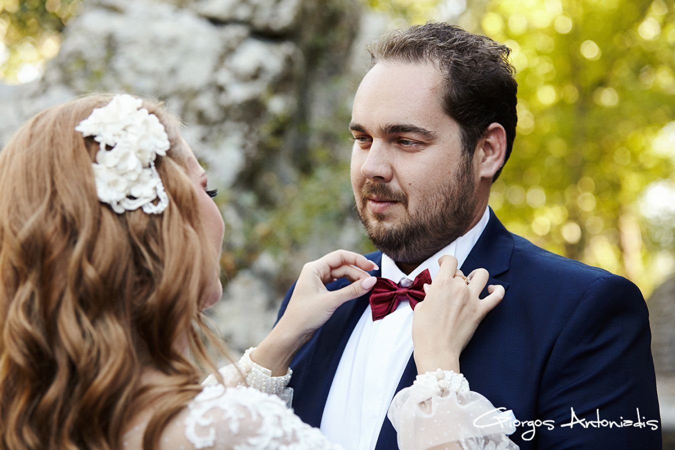 A couple dressed for a wedding, with the woman adjusting the man's bowtie outdoors, surrounded by trees and natural scenery.