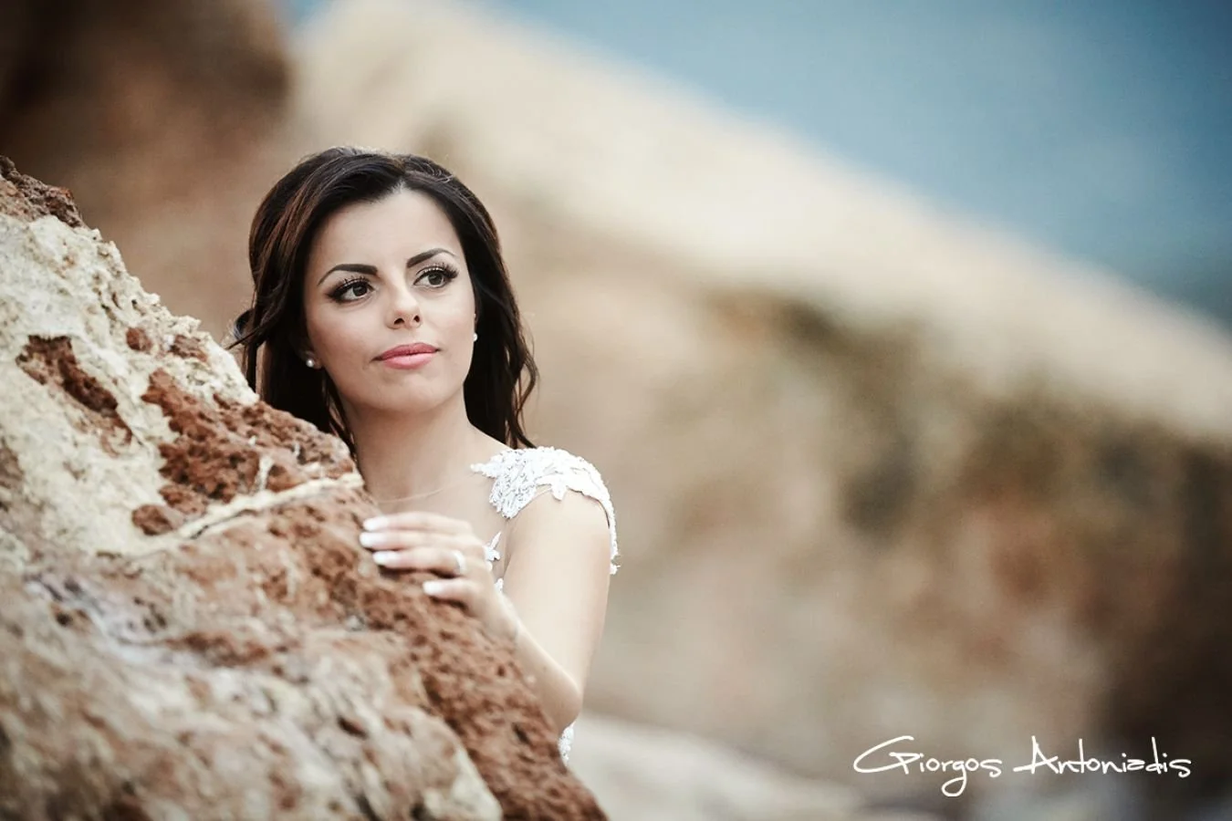 A woman with dark hair and makeup leaning on a rocky formation at the beach, wearing a white dress with lace details, with a blurred beach and sky in the background.