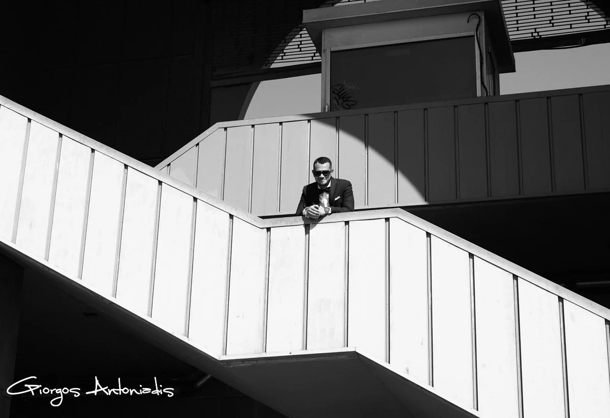 A man in a suit and sunglasses leaning on a white railing, on a modern outdoor staircase or balcony with shadows and geometric architectural elements.