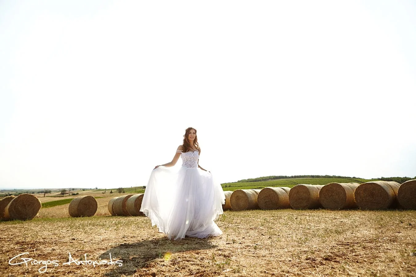 A woman in a white wedding dress standing in a field with hay bales, bright sky in the background.