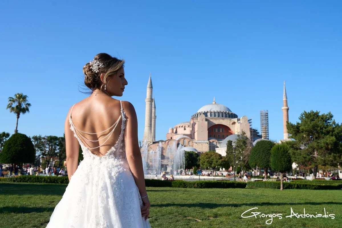 Bridal woman in white wedding dress standing in park with Hagia Sophia in the background, clear blue sky, and lush green trees.