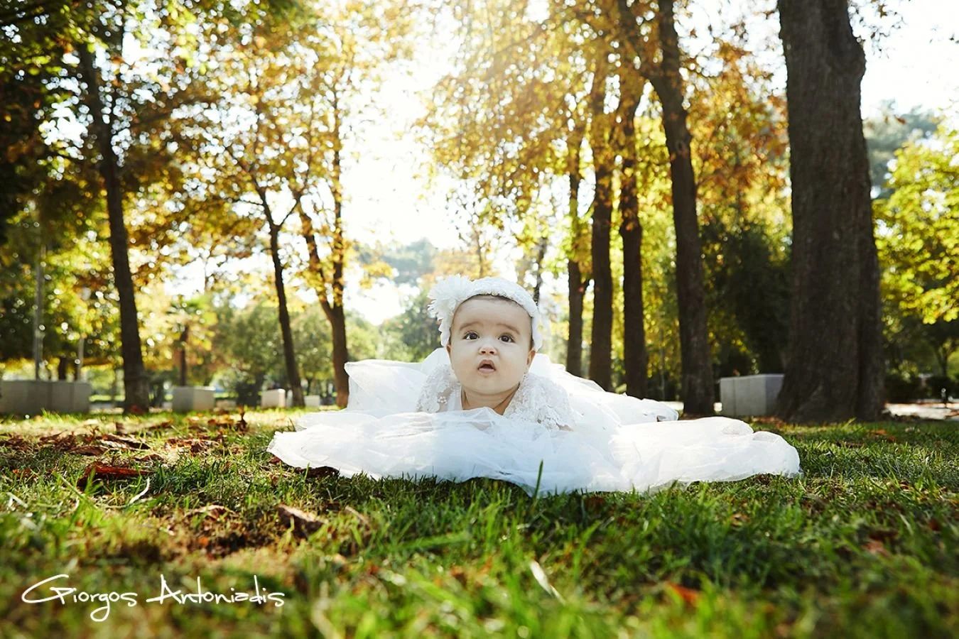 Baby girl in a white dress with a headband lying on her stomach on a grassy park with tall trees and sunlight in the background.