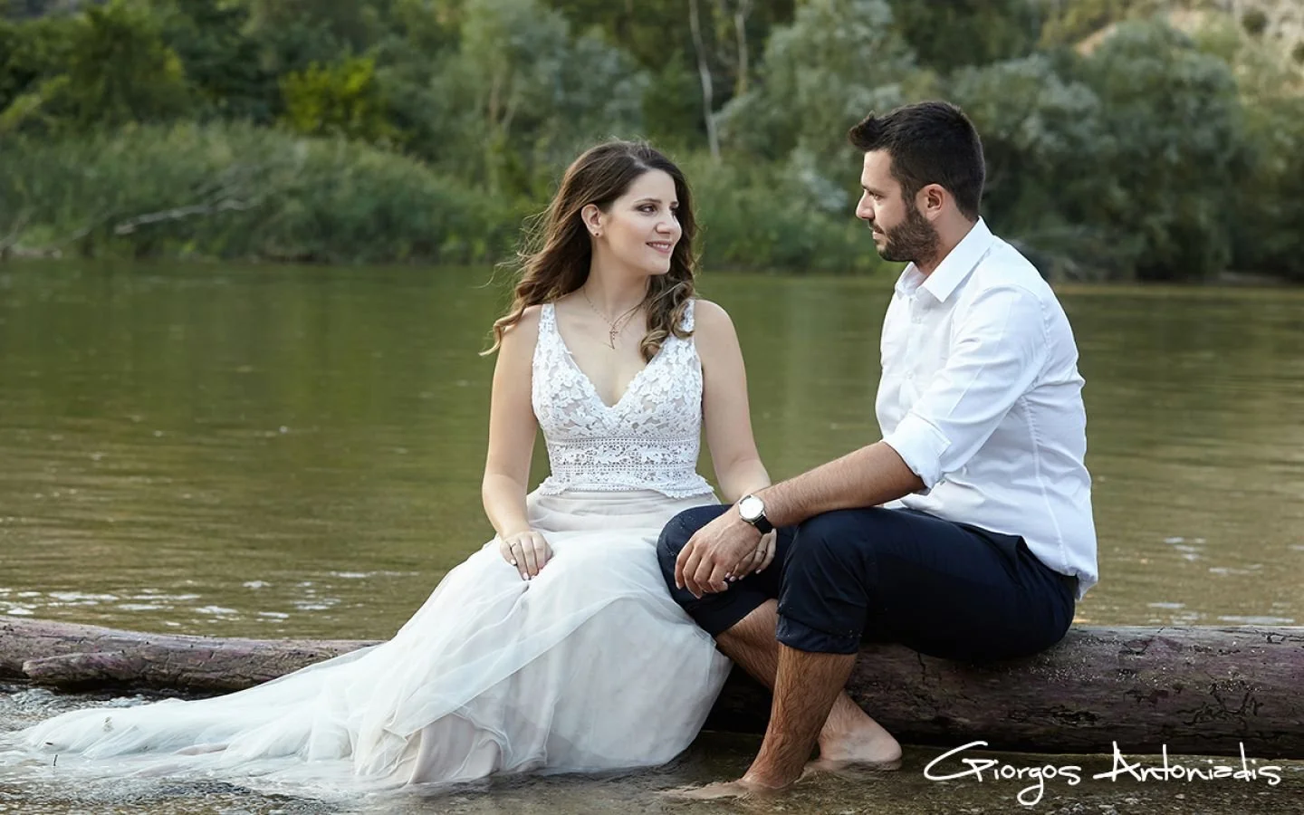 A couple sitting on a log in a river, looking at each other. The woman wears a white lace dress, and the man wears a white shirt and dark pants, both barefoot. Green trees are in the background.