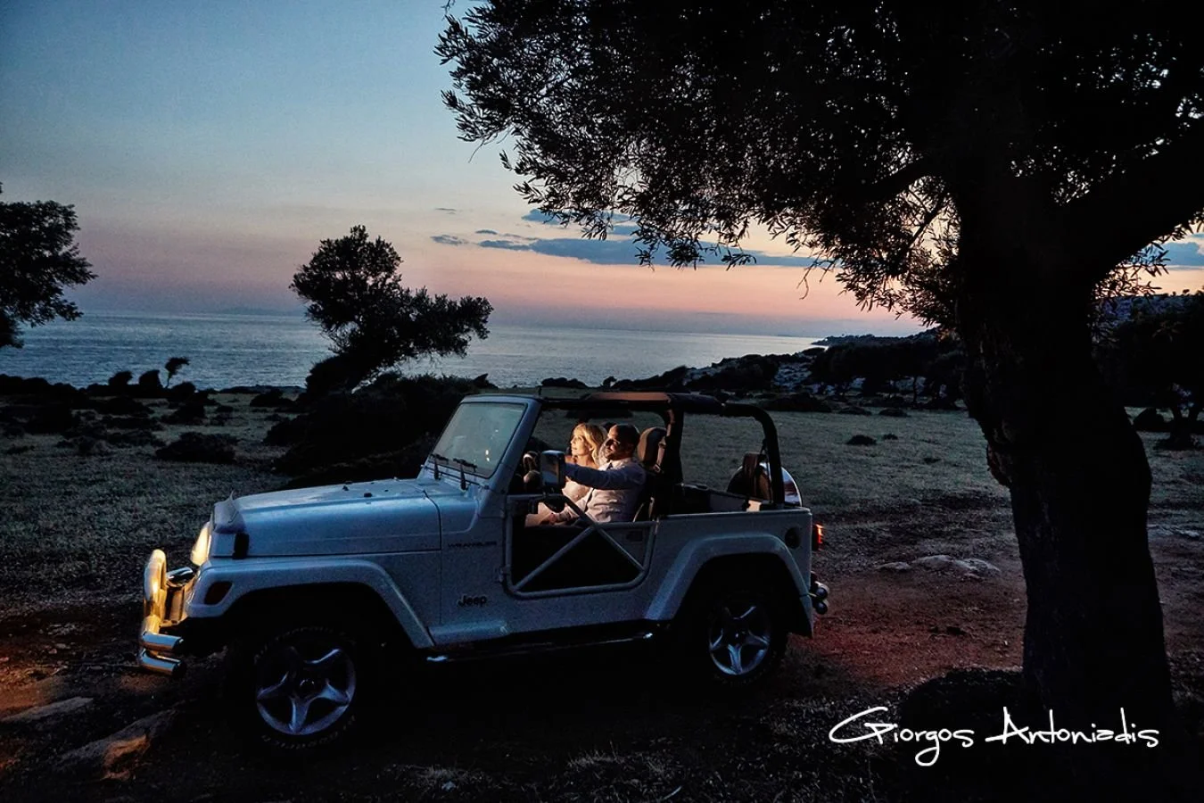 A white Jeep with two women inside, parked near the coast at sunset, with trees and rocky shoreline in the background.
